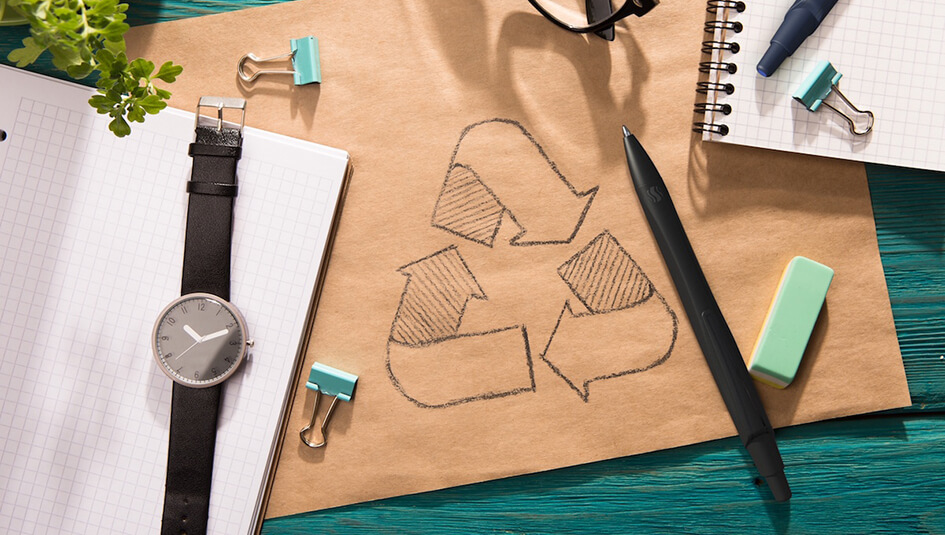 Recycling-Symbol auf Papier umgeben von Stift, Uhr und Büromaterialien, die nachhaltige Büroorganisation zeigen.