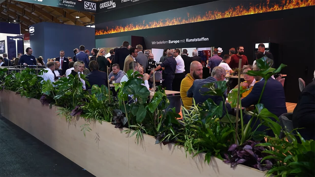 Menschen bei einer Kunststoffmesse in lebhafter Diskussion hinter grüner Pflanzendekoration.
