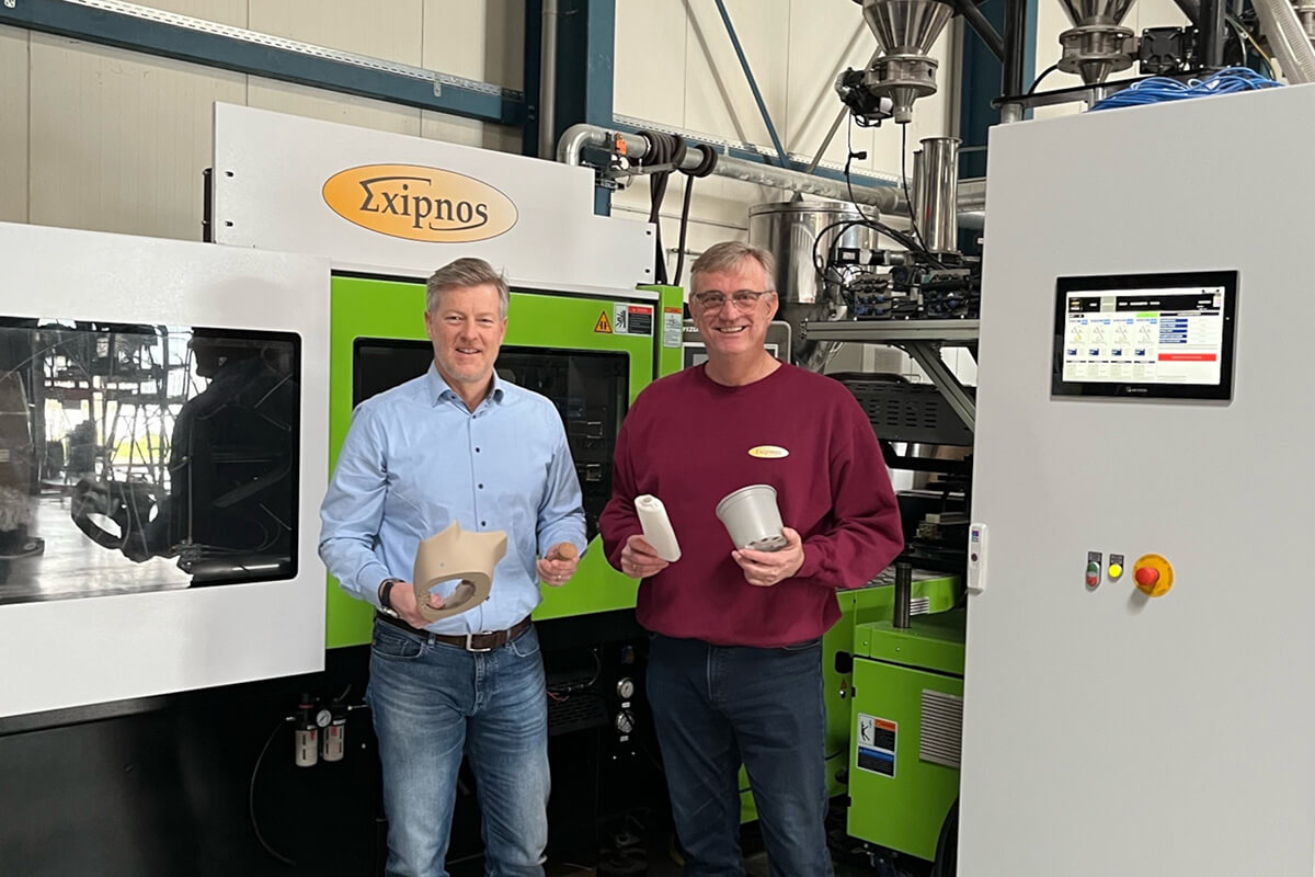 Zwei Männer präsentieren 3D-gedruckte Produkte vor einer Spritzgussmaschine in einer Fabrikumgebung.