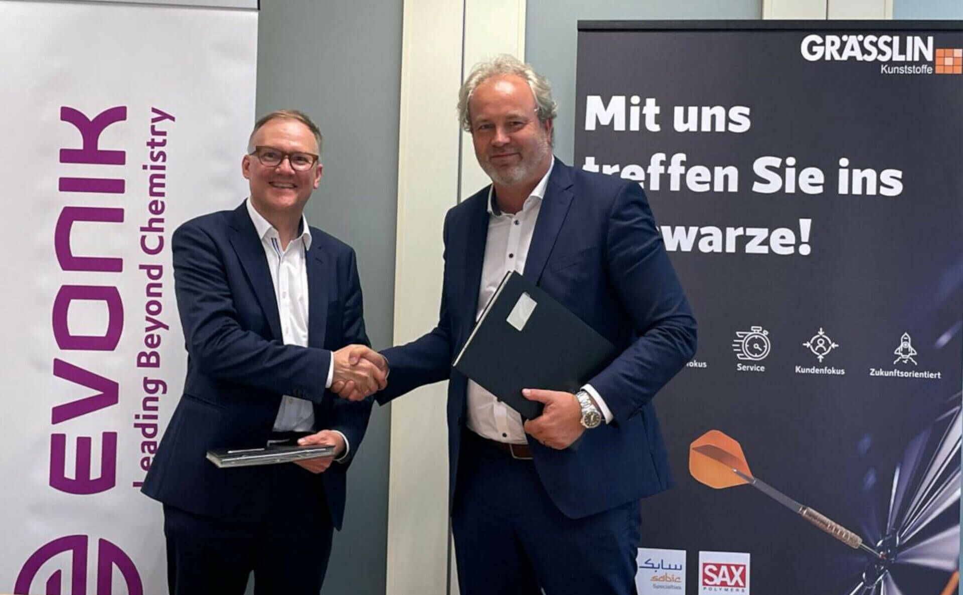 Zwei Männer in Anzügen schütteln sich die Hand vor Evonik- und Grässlin-Bannern in einem Geschäftsmeeting.