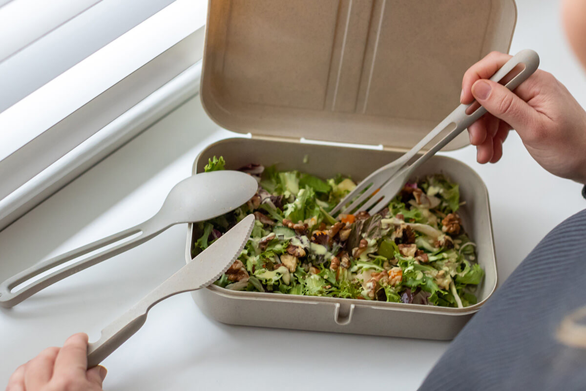 Frischer Salat in einer umweltfreundlichen Lunchbox mit Besteck auf einer weißen Oberfläche.