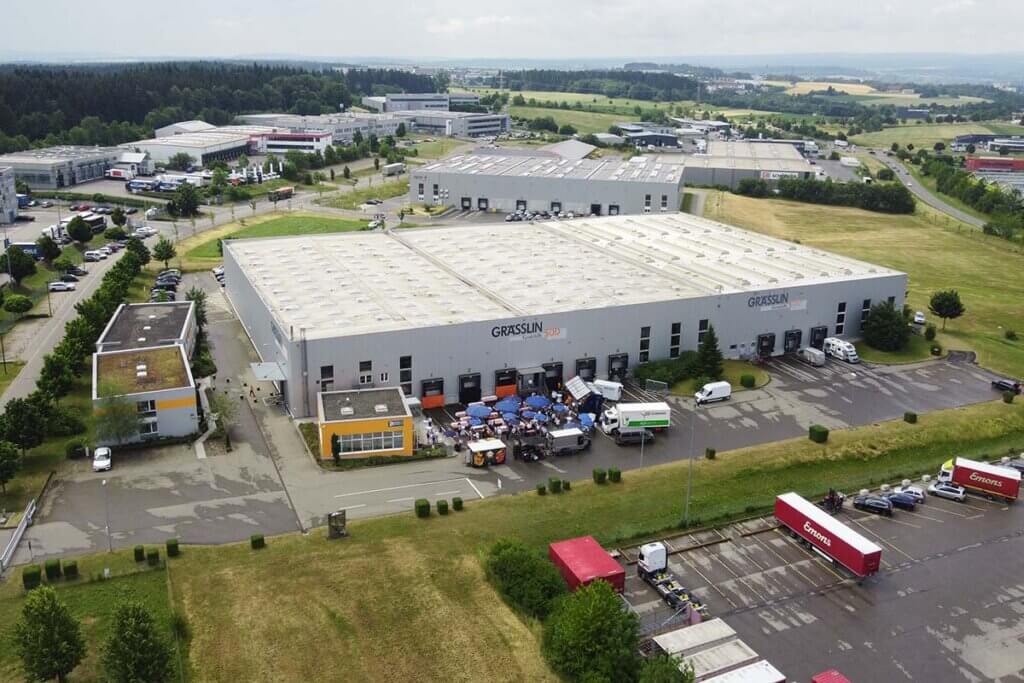 Industriegebäude mit LKWs und Parkplätzen aus der Luftperspektive in grüner Landschaft.