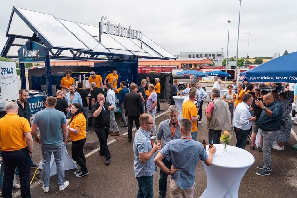 Menschen genießen ein Outdoor-Event mit Getränkestand und Gesprächsgruppen, entspannte Networking-Atmosphäre.