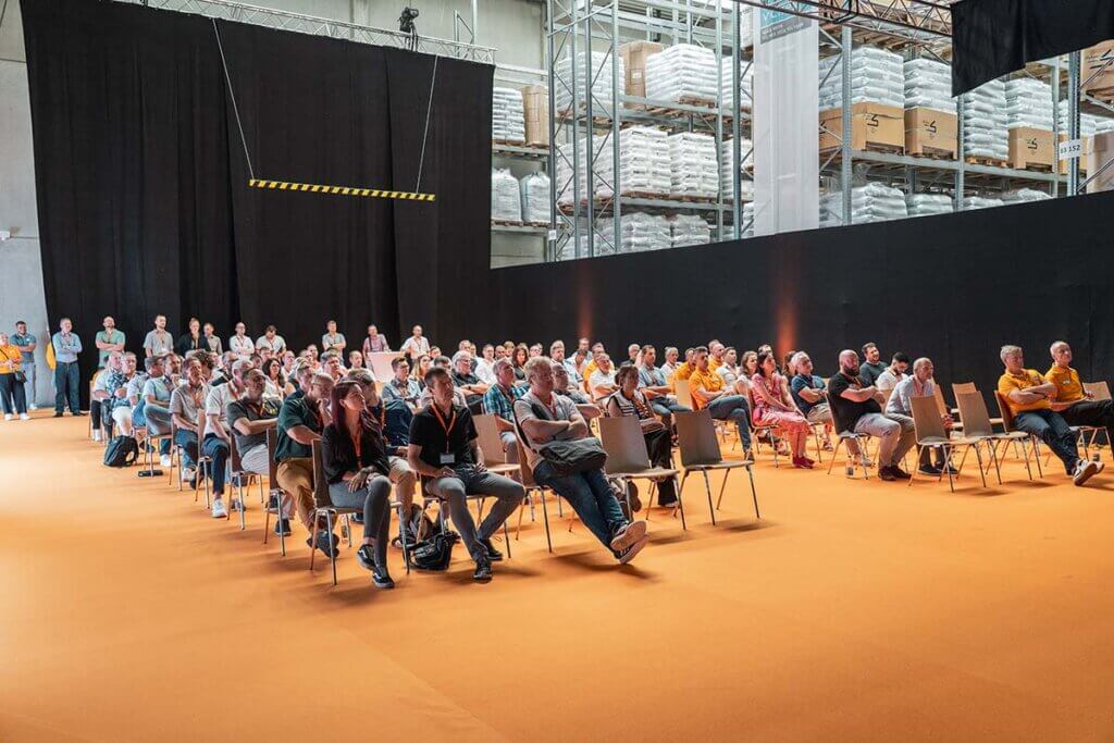 Publikum hört Vortrag bei Konferenz in Industriehalle, orangefarbener Boden, gefüllte Regale im Hintergrund.
