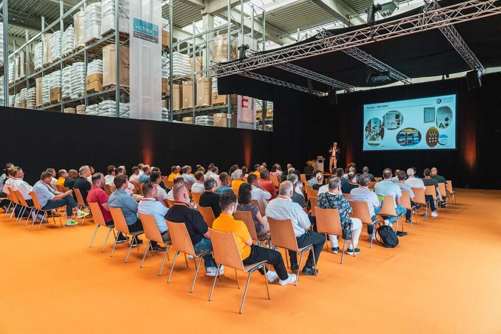 Konferenz in einem Lagerhaus mit Präsentation, Teilnehmer auf orangefarbenem Boden sitzen und zuhören.