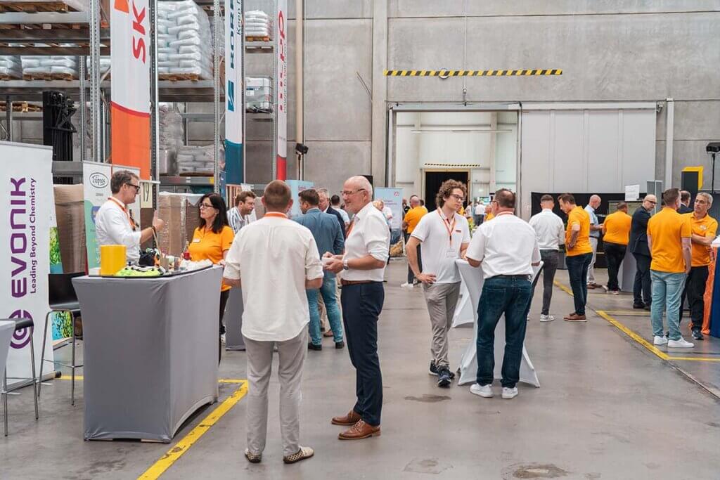 Menschen auf Networking-Event in Lagerhalle mit Infoständen und Gesprächen, Banner von Evonik und anderen Marken sichtbar.