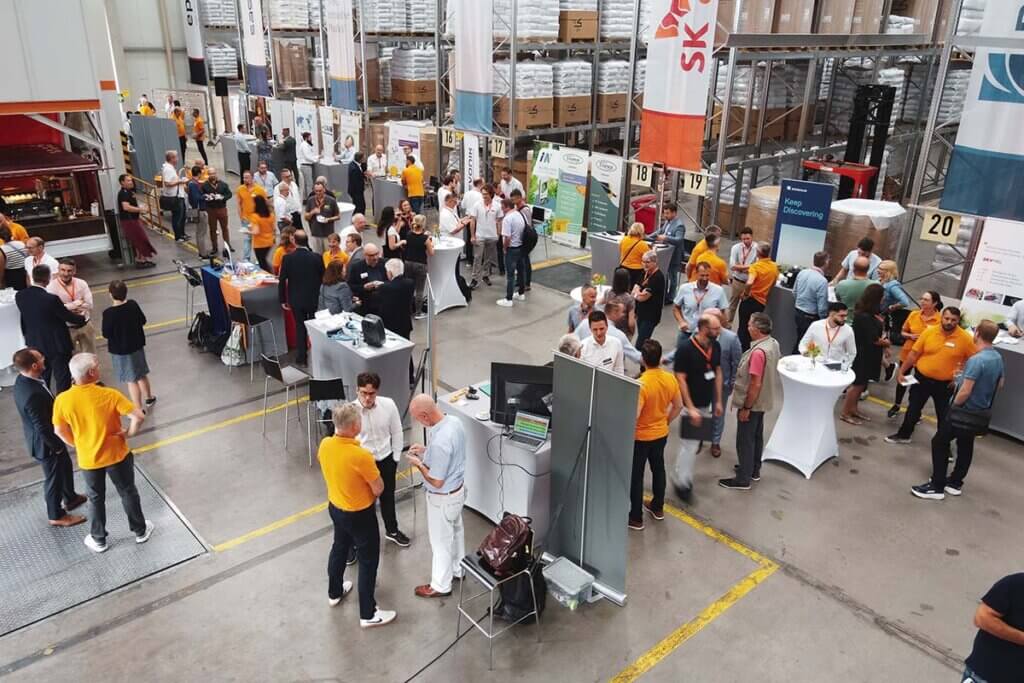 Konferenz in Lagerhalle: Menschen in Diskussionen, Infostände, Networking, lebendige Atmosphäre.