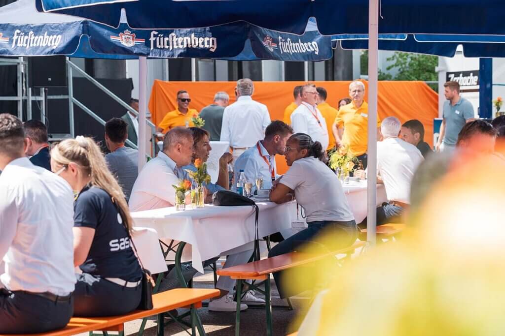 Menschen bei einem Firmenevent im Freien, Gespräche an Biertischen unter Fürstenberg-Sonnenschirmen.