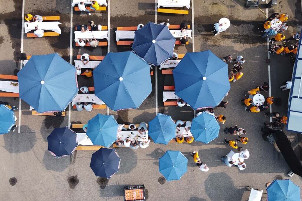 Draufsicht auf ein Outdoor-Event mit blauen Sonnenschirmen und Besuchern an Tischen auf einem Parkplatz.